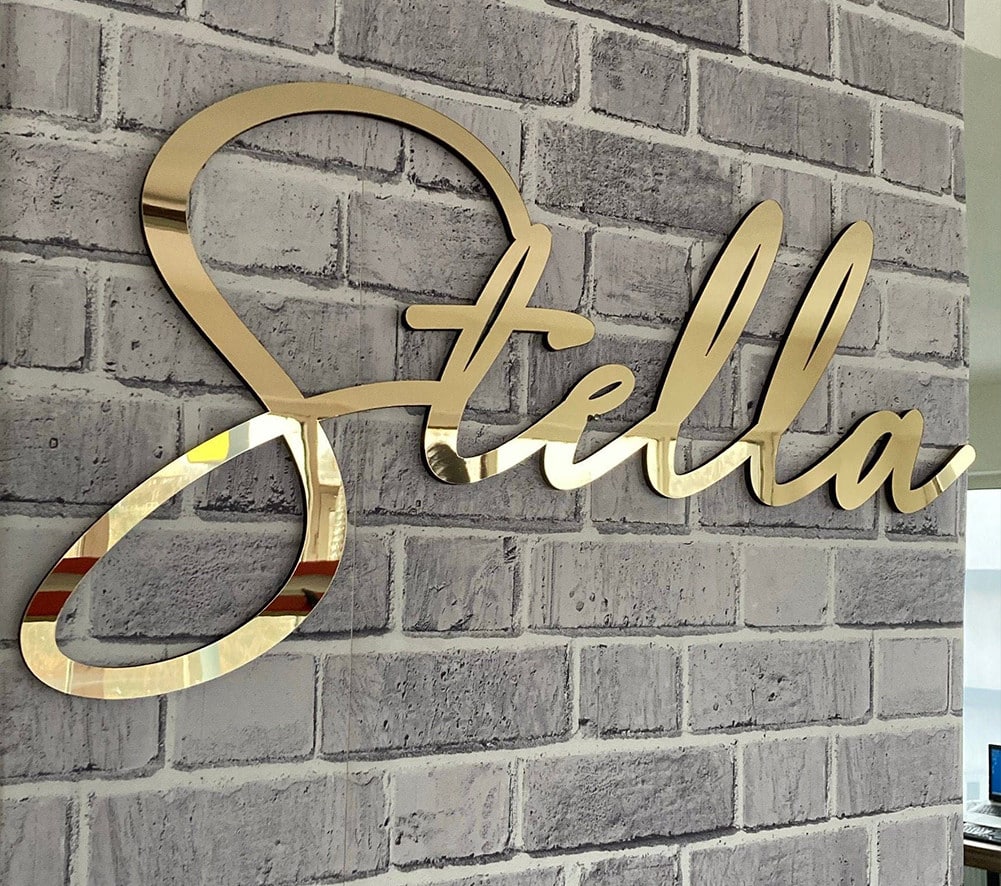 שלט קיר מוזהב עם הכיתוב "Stella" בכתב יד, המותקן על קיר לבנים אפור.