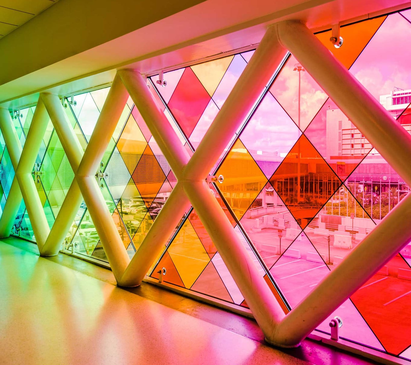 חלונות ויטראז' גיאומטריים צבעוניים עם דוגמאות יהלומים, הממוסגרים בקורות לבנות, משקיפים על אזור עירוני חיצוני עם בניינים ומדרכות.