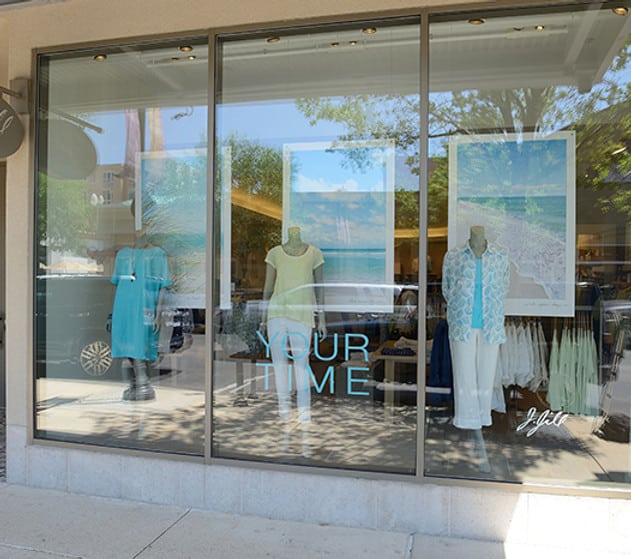 חלון ראווה בחזית החנות, בו מוצגים שלושה בובות ראווה לבושות בבגדים בצבעי תכלת ולבן, עם פוסטרים גדולים בנושא חוף הים והכיתוב "YOUR TIME" על הזכוכית.