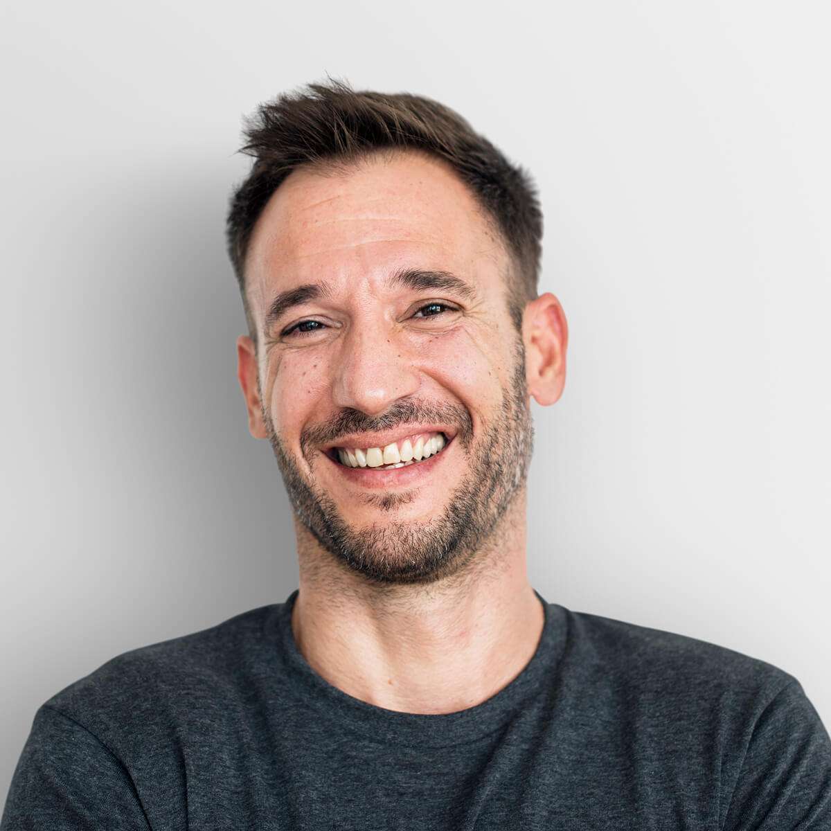 גבר עם שיער חום קצר וזקן מחייך למצלמה, לבוש בחולצת טריקו אפורה כהה, עומד על רקע בהיר וחלק.