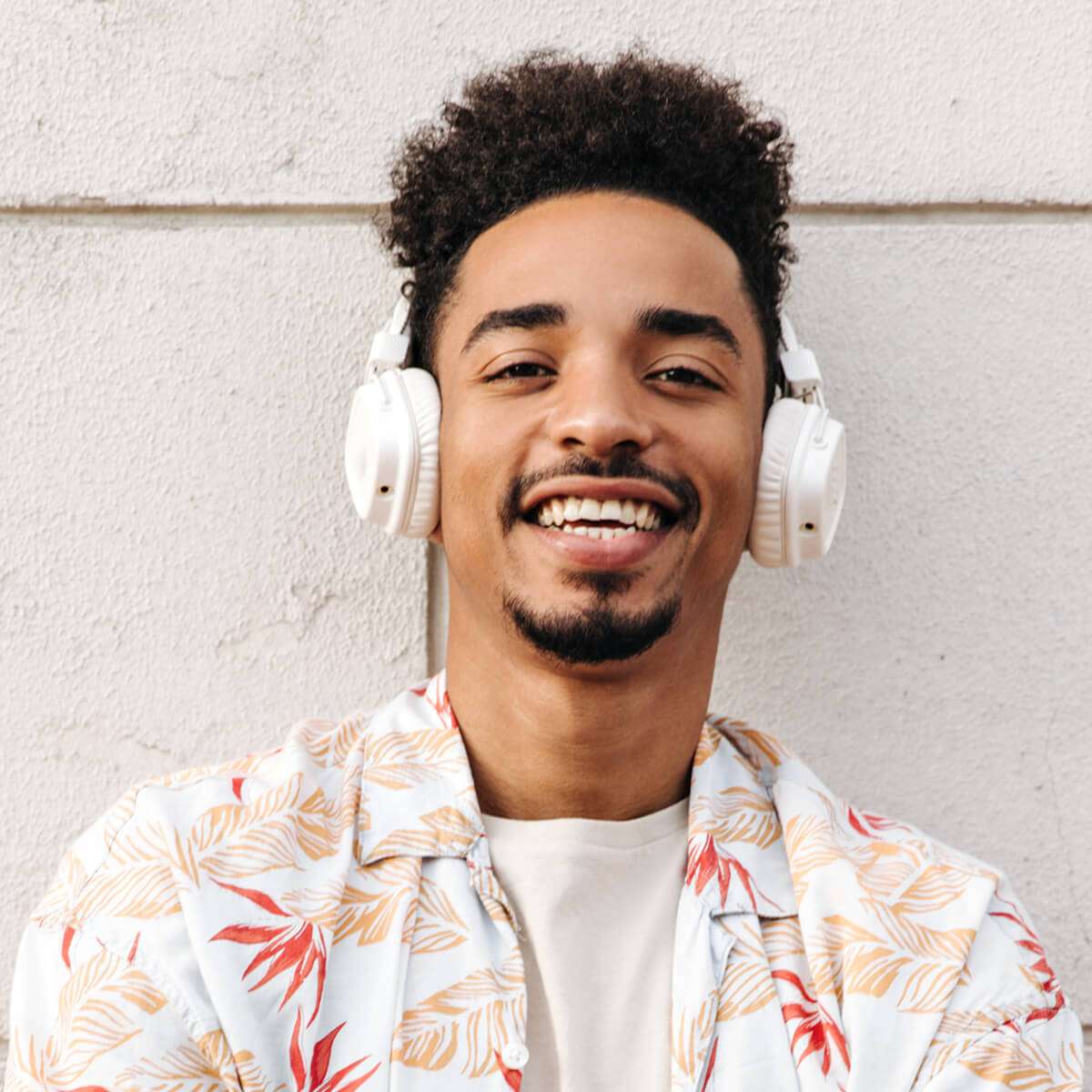 צעיר עם אוזניות וחולצה מעוטרת מחייך בעודו עומד מול קיר בהיר.